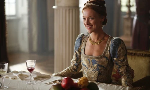 The character of Queen Elizabeth I sits smiling at a table