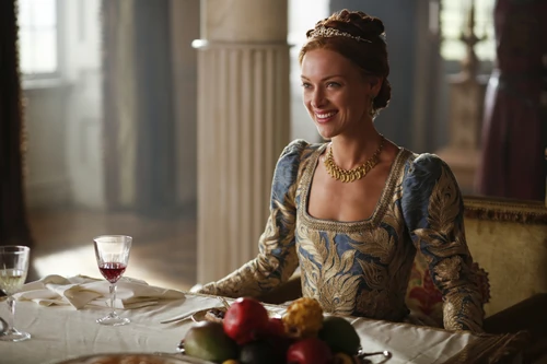 The character of Queen Elizabeth I sits smiling at a table