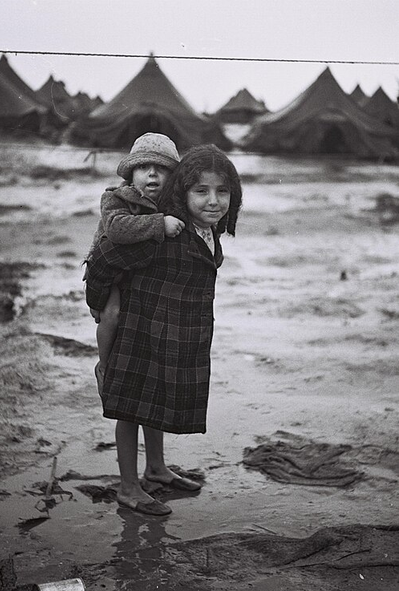 A young girl carries her younger brother on her back through a muddy field.